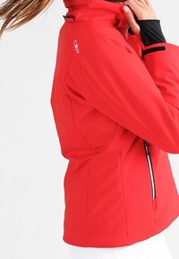 Rød jakke med et tekstureret fiskebenmønster, høj krave og sorte detaljer. Sidelås og logodetalje på skulderen.