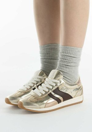 Voeten met grijze sokken en glanzende gouden sneakers met witte veters en bruine strepen, staand op een wit oppervlak.