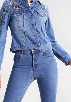 Kurtka jeansowa z haftem kwiatów i motyli, zestawiona z wysokim stanem niebieskich obcisłych jeansów na stojącej osobie.