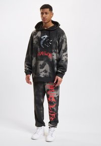 Felpa con cappuccio oversize nera e grigia con grafica frontale e testo rosso, abbinata a pantaloni coordinati con accenti di design rossi. Sneakers bianche.