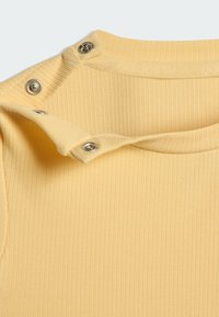 Żółta prążkowana tkanina z okrągłym dekoltem ozdobionym srebrnymi zatrzaskami na lewym ramieniu. Gładka faktura, szczegół z bliska.