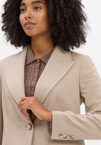 Vrouw met krullend haar die een knoop aanpast op een beige blazer, gedragen over een bruine kabeltrui met knopen, tegen een witte achtergrond.