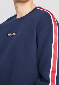 Sweatshirt marine à col rond, avec un patch noir "HOLLISTER" et des accents rayés rouges et blancs sur les manches. Texture de tissu douce.