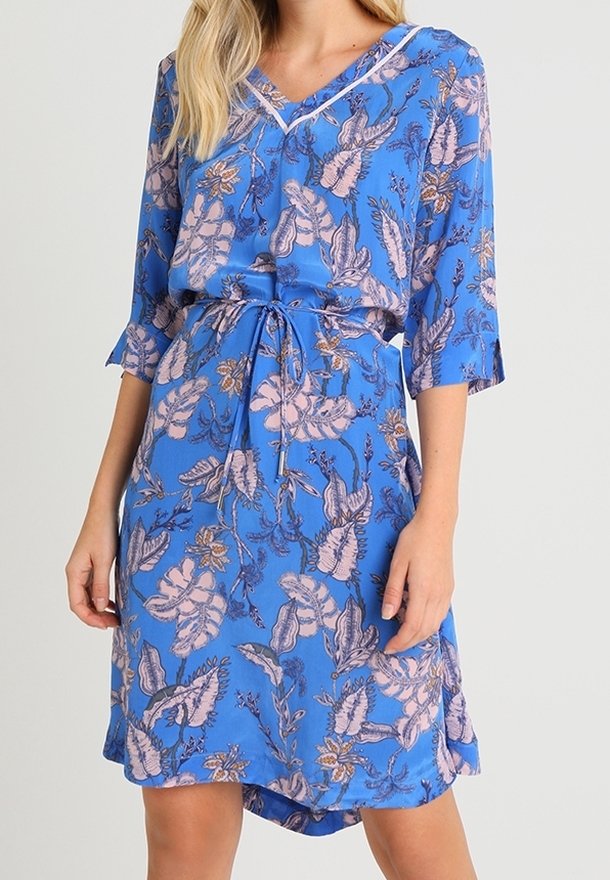 Femme portant une robe bleue longueur genoux avec motifs floraux et feuillus roses, manches trois-quarts, col en V et ceinture à nouer à la taille.