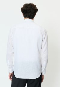 Moški v dolgo rokavčni beli laneni srajci in črnih hlačah, stoji obrnjeni stran proti enostavni svetli podlagi.