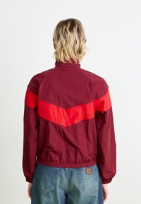 Bordeaux lett jakke med rød sveitsers design, høy krage og elastiske mansjetter. Glatt tekstur og løs passform, kombinert med blå jeans.