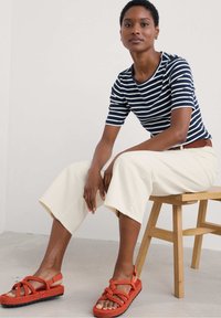 Striped navy and white ribbed shirt, cream wide-leg pants, and orange knotted sandals. Model seated on a wooden chair. Minimalist background.