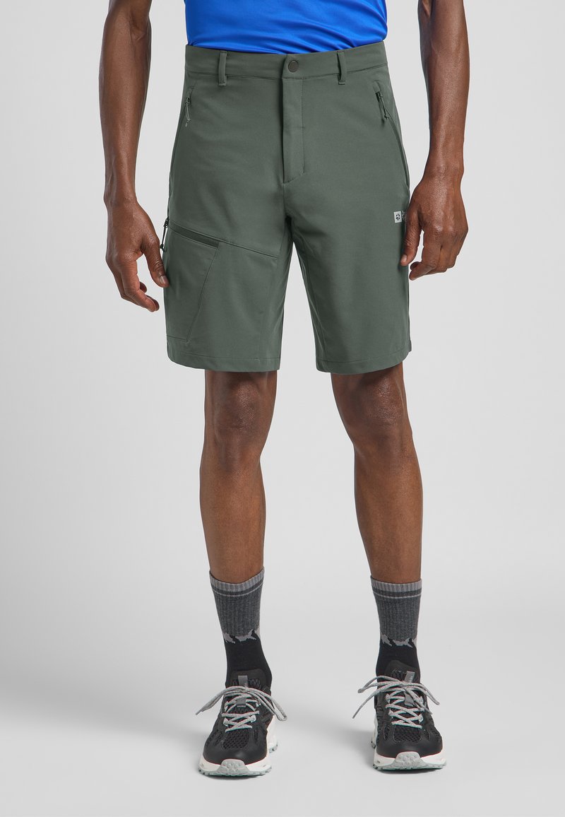 Shorts verts en tissu extensible et respirant. Les caractéristiques comprennent plusieurs poches et une fermeture éclair. Associés à des chaussures noires et des chaussettes grises.