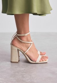Beige Absatzsandalen mit einem strukturierten Krokodilmuster, ausgestattet mit dünnen, über Kreuz verlaufenden Riemen und einem klobigen quadratischen Absatz.