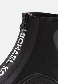 Detailní záběr na černou pletenou kotníkovou botu s vertikálním zipem, bílým textem "MICHAEL KORS" a kulatým táhlem zipu s logem MK.