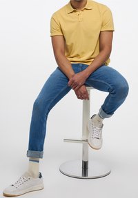 Keltainen polo-paita, siniset kapeat farkut, valkoiset tennarit meriharmailla yksityiskohdilla ja vaaleat sukat. Istuu tuolilla jalat ristissä.