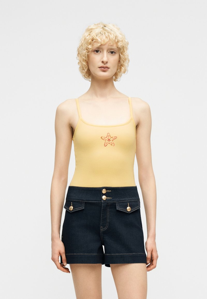 Γυναίκα με σγουρά ξανθά μαλλιά που φοράει κίτρινη αμάνικη μπλούζα με σχέδιο αστερία και σκούρο jean σορτς με χρυσά κουμπιά.