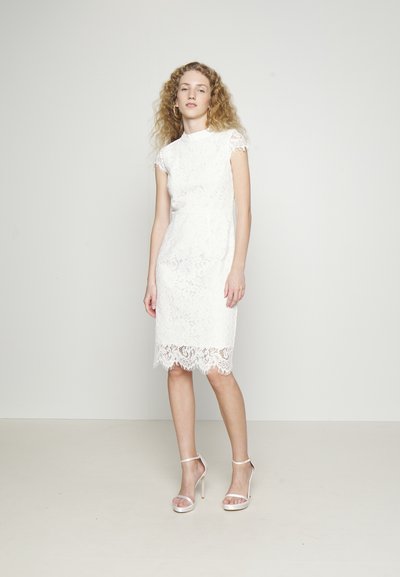 Λευκό δαντελένιο φόρεμα με κοντά μανίκια, εφαρμοστή σιλουέτα, θηλυκή περίτεχνη ραφή και περίπλοκα floral σχέδια. Συνδυάζεται με λεπτές σανδάλες με λουριά.