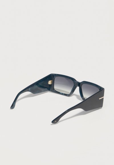 Σκούρα μπλε υπερμεγέθη τετράγωνα γυαλιά ηλίου με παχιά πλαίσια και βαθμιαία σκουρότερα φακούς, τοποθετημένα πάνω σε ανοιχτό γκρίζο επιφάνεια.