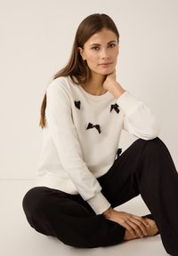 White sweatshirt with black bow details, round neckline, and ribbed cuffs, paired with loose black pants. Smooth fabric texture.