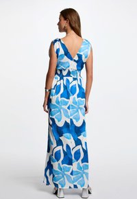Abito lungo in tessuto fluido con un motivo floreale astratto blu. Design a V sulla schiena, vita arricciata e maniche corte. Scarpe argento visibili.