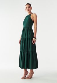 Zeleni maxi obleka z visokim ovratnikom, večplastno krilo in brezrokavnim dizajnom. Mehka tekstura, tekoča tkanina, kombinirana s telovniki v nude barvi.