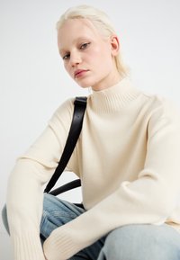 Helle beige Strickpullover mit hohem Kragen und gerippter Textur; getragen mit blauen Jeans. Ein schwarzer Riemen einer Tasche liegt auf der Schulter.