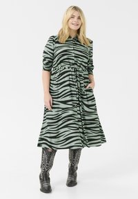 Abito verde a strisce a zebra con bottoni frontali, vita cinte e tasche, abbinato a stivali da cowboy neri ricamati.
