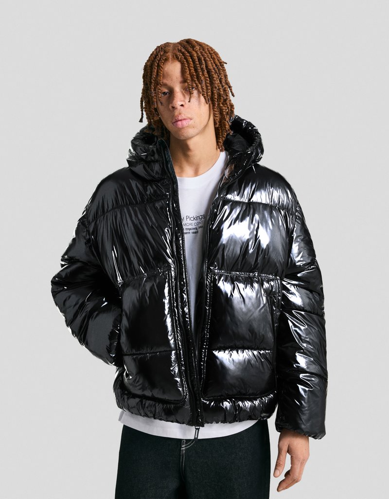 Black puffer jacket made of shiny, quilted material; features a hood and two front pockets; worn over a white shirt and dark jeans.