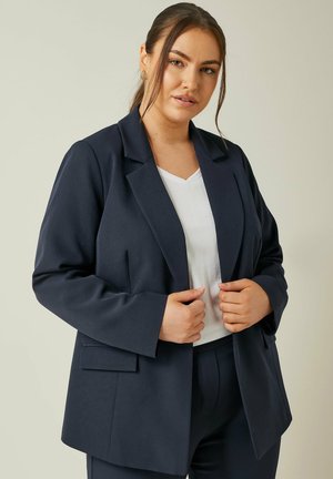 Blazer marine avec une texture lisse, des revers crantés, des manches longues et des poches latérales, porté par-dessus un haut blanc à encolure en V.