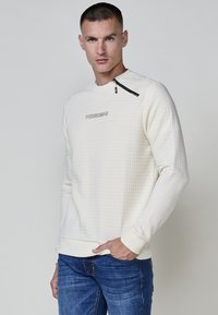 Sudadera blanca acolchada con un acento de cremallera en el hombro, puños de canalé y un logo en el centro. Combinada con jeans azules.