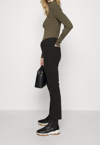 Jeans negros de corte ajustado, alta cintura y bolsillos frontales, combinados con una blusa de canalé color oliva y botas negras de tobillo con suela blanca.