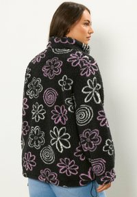 Czarna polarowa kurtka z wielokolorowymi kwiatowymi i spiralnymi wzorami w białym i purpurowym kolorze. Wysoka stójka i luźny krój, z bocznym sznurkiem do regulacji.
