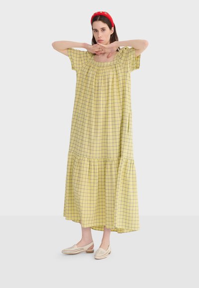 Femme portant une robe midi ample à carreaux jaunes, des ballerines tissées crème et un bandeau rouge, debout les bras levés et les mains croisées.