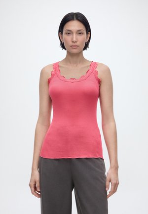 Γυναίκα με κοντά σκούρα μαλλιά, που φοράει εφαρμοστή ροζ κοραλί φανέλα με δαντέλα και χαλαρά σκούρα γκρι παντελόνια, στέκεται μπροστά σε απλό φόντο.