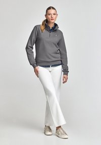 Grå zip-tröja, vita utsvängda byxor, beige sneakers. Skjorta i denim lager på lager under. Modell med långt hår står framför ett neutralt bakgrund.