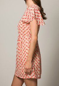 Kreemi mekko, jossa on punaisia kirsikkakuoseja, lyhyet liehuvat hihat ja istuva vyötärö. Pehmeä kangas, jossa on kevyt tekstuuri. Keskipituus reiden yläosaan.