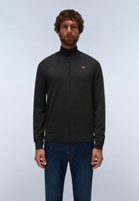 Man with curly hair and beard wearing a dark gray zip-up sweater and blue jeans, standing against a plain light background.
