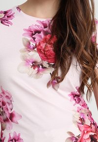 Top con estampado floral que presenta grandes flores rosas y moradas sobre un fondo rosa claro, con un diseño de cuello redondo y una textura de tela suave.