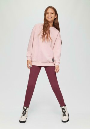 Roze sweatshirt met verlaagde schouders, gecombineerd met bordeauxrode leggings. Het model draagt witte veterschoenen met zwarte accenten. Eenvoudige, casual outfit.