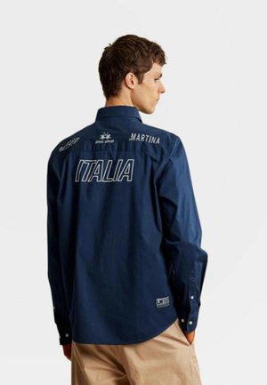 Man draagt een donkerblauw shirt met lange mouwen met de tekst "ITALIA" en "MARTINA" op de rug, gecombineerd met een beige broek, met zijn rug naar de camera.