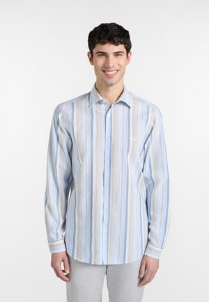 Junger Mann lächelt, trägt ein hellblau-weiß-beiges Hemd mit vertikalen Streifen, Knopfleiste und langen Ärmeln sowie eine hellgraue Hose.