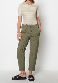 Pruhované béžové tričko s krátkým rukávem, olivově zelené kalhoty s elastickým pasem a šnůrkou, a černé sandály na ploché podrážce se šněrováním.