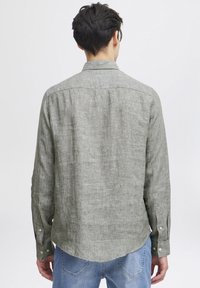 Camicia button-up verde in lino con un retro a yoke, orlo arrotondato e maniche lunghe, abbinata a jeans azzurri chiari. Tessuto morbido e design casual.