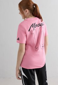 Punapea punase juustega tüdruk, kellel on hobusesaba, kannab roosa Messi 10 särki ja musti sportpükse, seistes ühetaolise valge seina vastas.