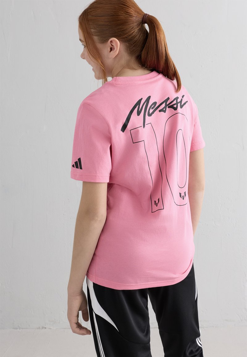 Punapea punase juustega tüdruk, kellel on hobusesaba, kannab roosa Messi 10 särki ja musti sportpükse, seistes ühetaolise valge seina vastas.