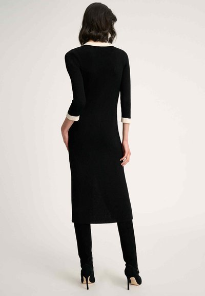 Robe en tricot côtelé noire avec des manches trois-quarts et un col crémeux contrastant, atteignant le mollet, associée à des bottes à talons aiguilles noires.