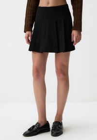Zwarte geplooide rok met een aansluitende taille, gemaakt van een gladde stof. Heeft een uitlopende silhuet die boven de knieën valt, gecombineerd met zwarte loafers.