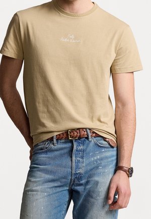 Homme portant un t-shirt beige Polo Ralph Lauren, une ceinture en cuir marron tressée, un jean bleu et une montre-bracelet en cuir marron.