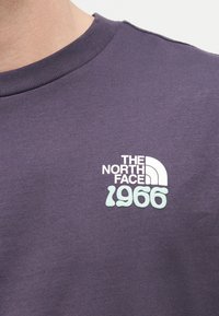 Κοντινή λήψη μοβ μπλουζάκι με στρογγυλή λαιμόκοψη και λευκό και ανοιχτό μπλε λογότυπο «The North Face 1966» στο στήθος.