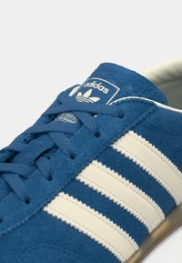 Μπλε σουέτ αθλητικό παπούτσι με λευκό σχέδιο τριών ρίγων, στρογγυλά κορδόνια και επενδεδυμένο γιακά που φέρει το λογότυπο της Adidas στη γλώσσα.