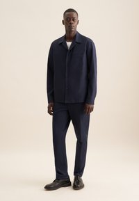 Camicetta blu navy, con bottoni, dotata di una tasca singola, abbinata a pantaloni su misura coordinati e scarpe nere stringate su uno sfondo chiaro.