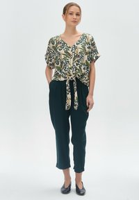 Bluzka z krótkim rękawem w kwiatowy wzór, z wiązaniem z przodu w kolorze kremowym, zielonym i granatowym. Spodnie w ciemnozielonym kolorze z podwiniętymi mankietami w zestawieniu z czarnymi balerinami.