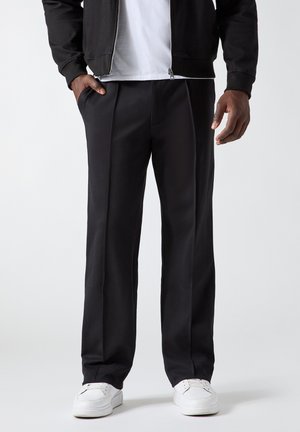 Homme portant un pantalon noir droit avec des poches latérales, un t-shirt blanc, une veste noire zippée et des baskets blanches sur fond blanc.