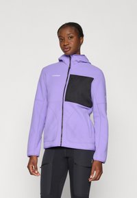 Person wearing a purple fleece jacket with a black chest pocket and black athletic leggings, standing against a plain light background.
